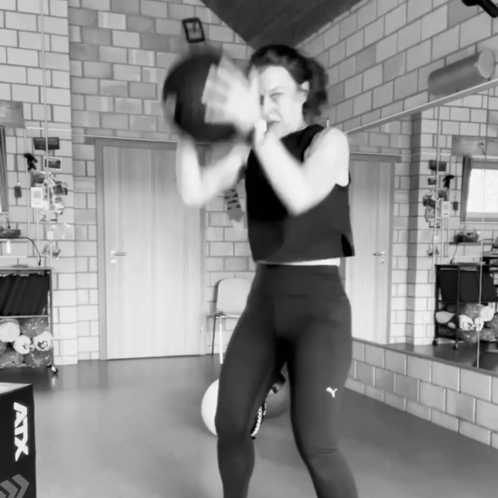 Das Bild zeigt Jo Angele hält einen Ball mit beiden Händen fest und ist dabei, ihn auf den Boden zu schmeissen. Sie führt die Übung "Slam Ball" aus. 